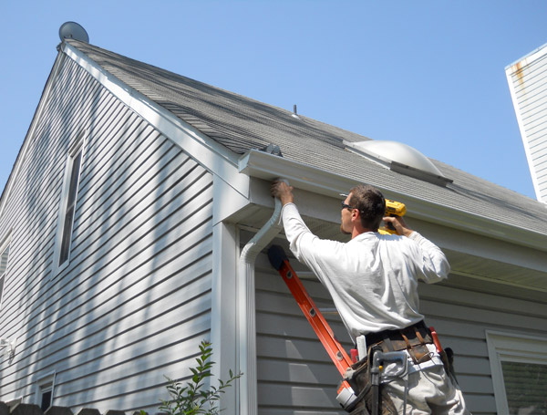 Supreme Restorations installer mounting a new white aluminum seamless gutter system on a suburban home